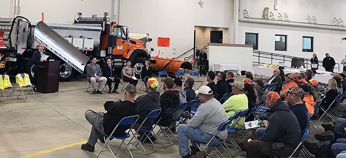 Workers sitting in folding chairs, facing the direction of a person reading from a podium. A snowplow is behind the person at the podium.